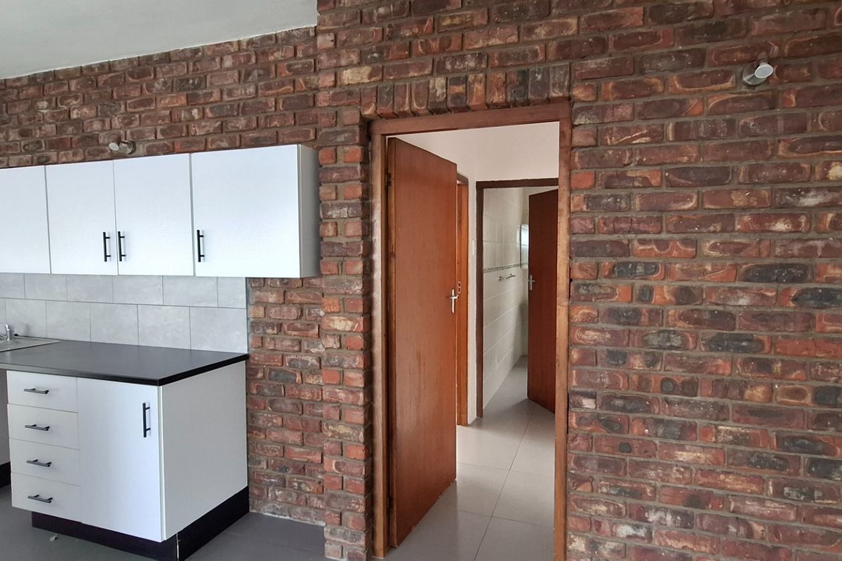 A kitchen area featuring white cabinetry, dark countertops, and a distinctive exposed brick feature wall, leading into a hallway with wooden doors.