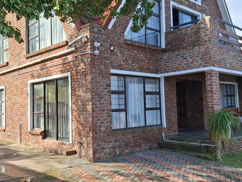 A multi-story face-brick house featuring a covered patio, balcony, and paved driveway area with large windows and a pitched roof design.