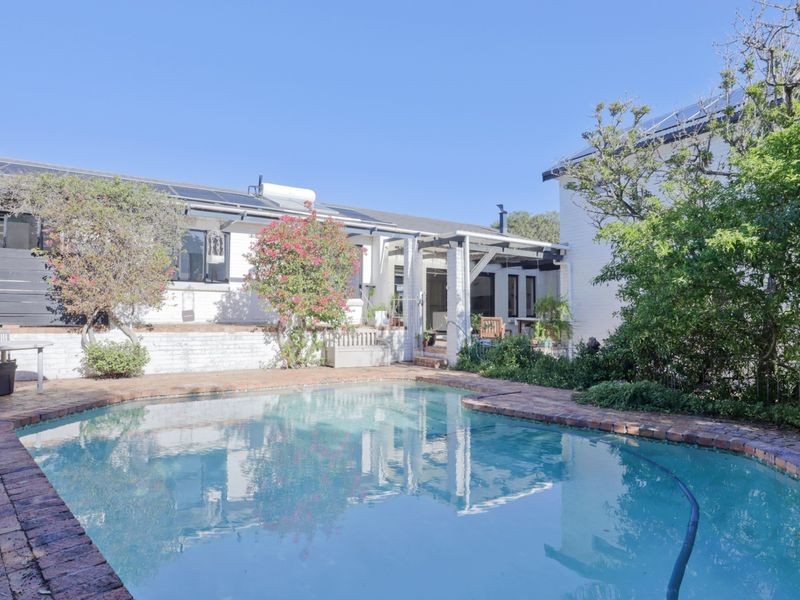 A well-maintained outdoor pool area featuring brick coping, a covered patio space, and solar panels installed on the roof of the main dwelling.