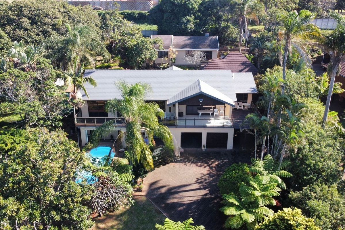 Aerial view of a residential property featuring a paved driveway, double garage, elevated balcony with glass balustrades, and a swimming pool set in lush gardens.