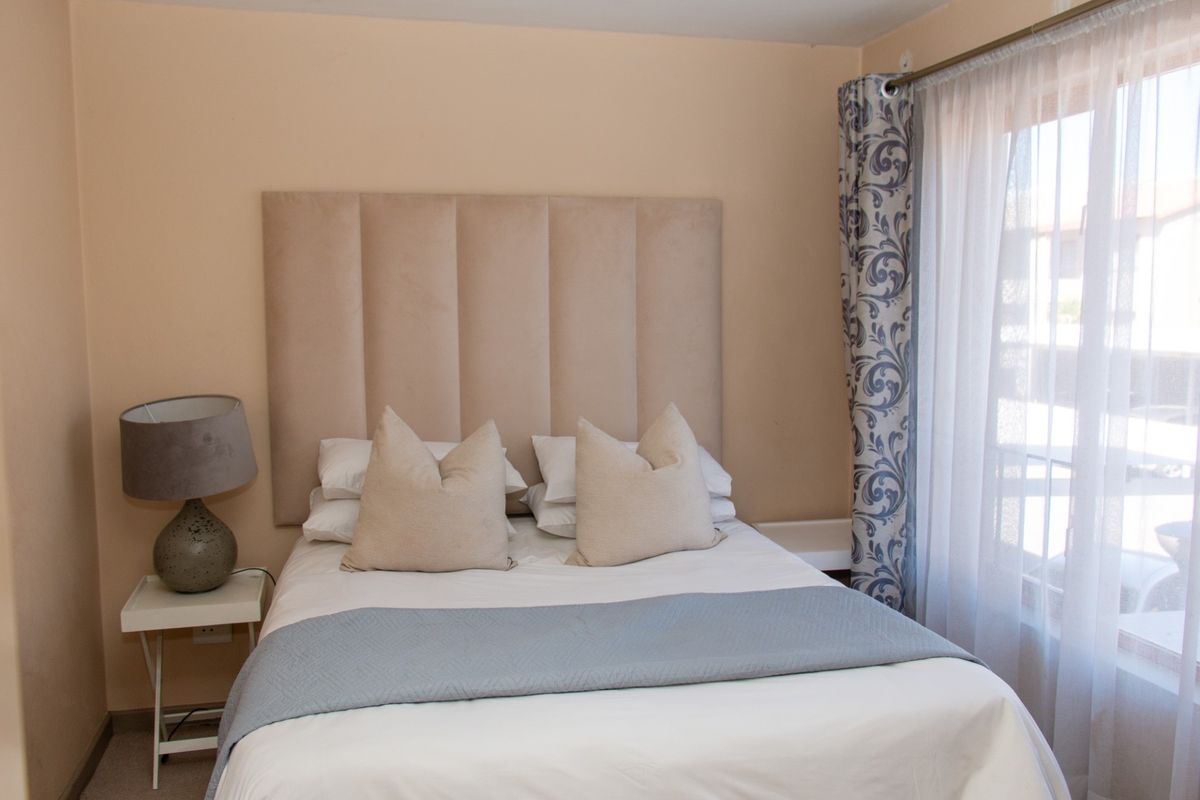 A cozy bedroom featuring neutral wall tones, a built-in headboard, and natural light provided by a window with patterned curtains.