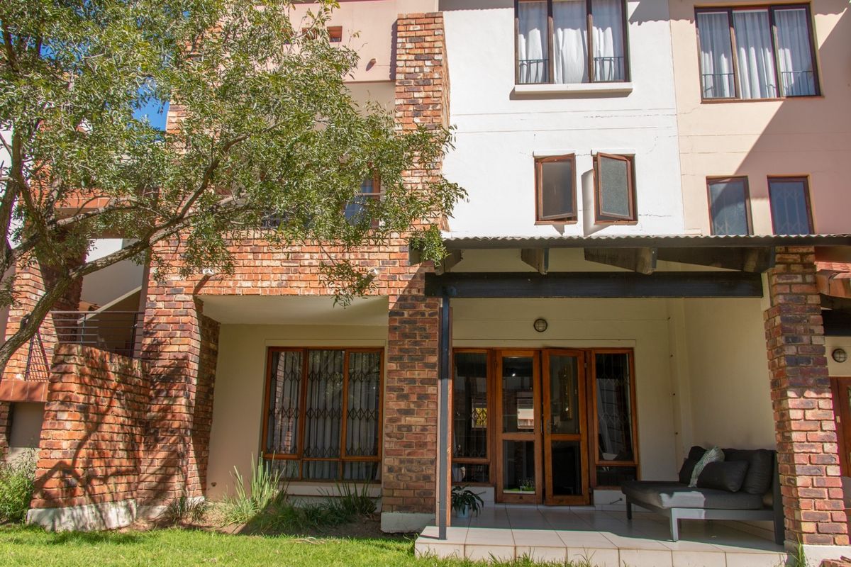 A multi-story residential exterior featuring exposed brickwork, a covered patio area, and tiled flooring, set against a small garden space.