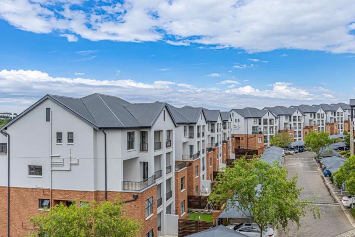 An aerial view of a modern apartment complex featuring multi-story buildings with pitched metal roofs, brick and plaster facades, and paved parking areas.