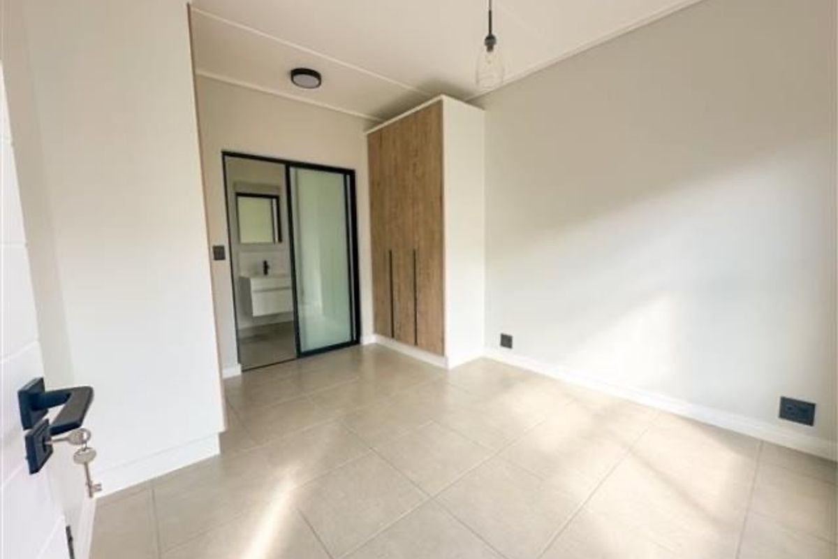 A bright, modern bedroom featuring neutral floor tiles, built-in wooden cabinetry, and an en-suite bathroom with a sliding glass door.