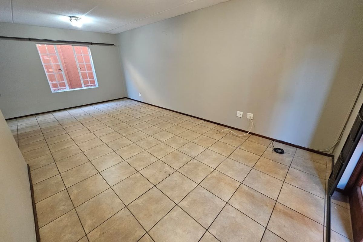 A spacious, empty living area featuring neutral wall paint, ceramic floor tiles, and a window fitted with security burglar bars.