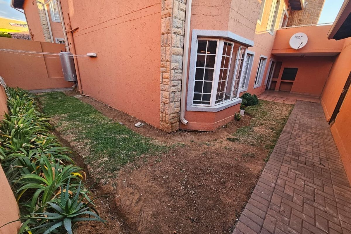 Rear exterior view of the townhouse featuring a paved walkway, bay window, stone cladding accents, and a small garden area with an external geyser.
