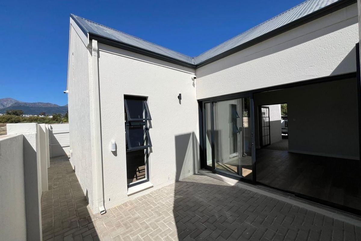 A modern rear exterior view featuring a paved patio area, sliding glass doors, and clean white rendered walls under a metal roof.