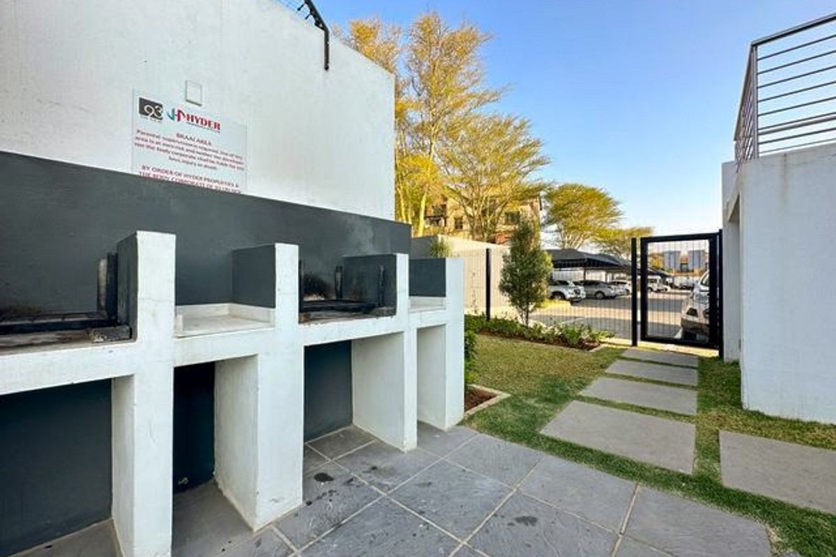 Outdoor communal braai area featuring built-in masonry barbecue stations, paved flooring, and access to the complex parking area.
