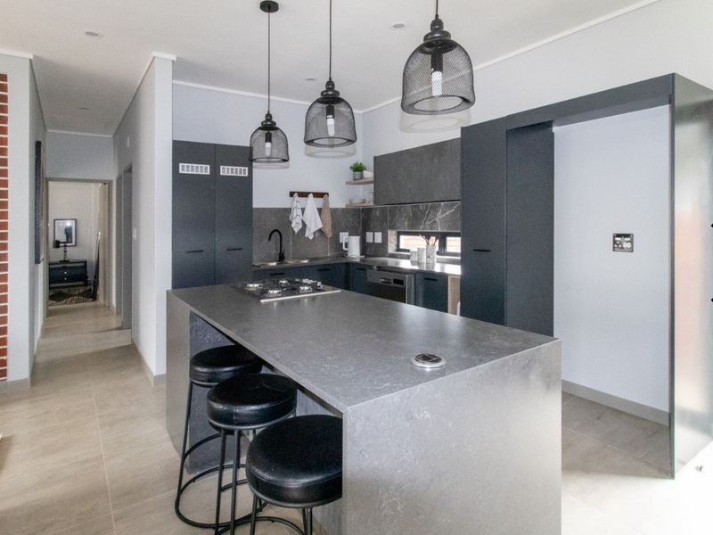 Modern kitchen featuring a central island with built-in hob, sleek dark cabinetry, stone countertops, and contemporary pendant lighting in an open-plan layout.