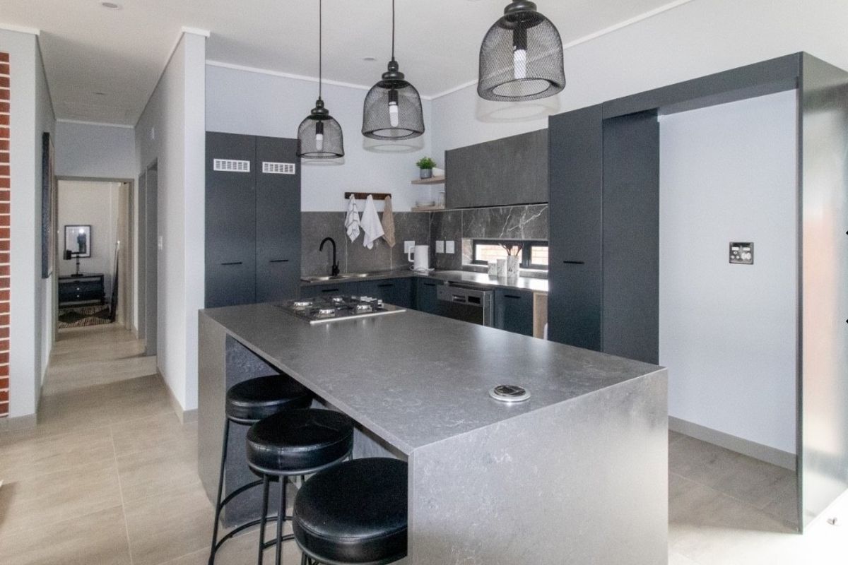Modern kitchen featuring a central island with built-in hob, sleek dark cabinetry, stone countertops, and contemporary pendant lighting in an open-plan layout.