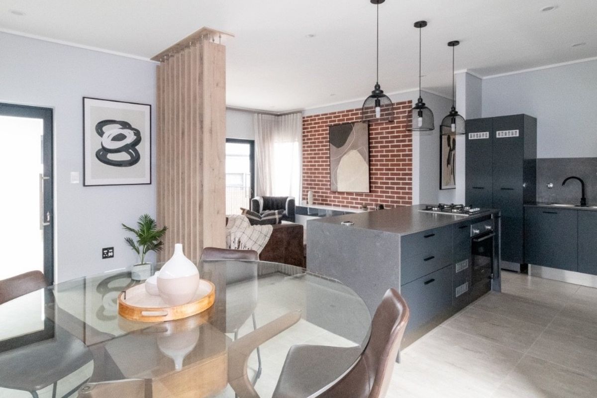 Modern open-plan living area featuring a sleek kitchen island, exposed brick accent wall, timber slat divider, and contemporary pendant lighting throughout the space.