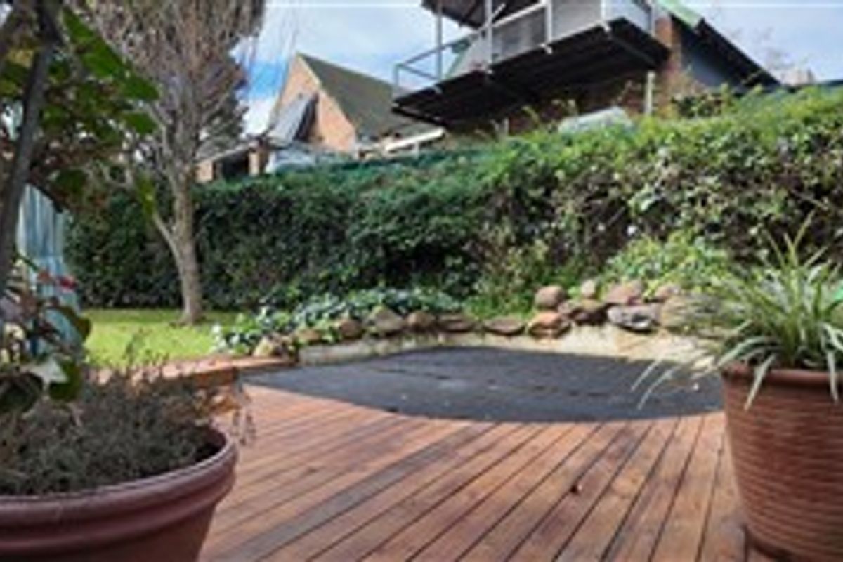A rear garden area featuring a timber deck, a covered pool, and tiered landscaping with established greenery and stone retaining elements.