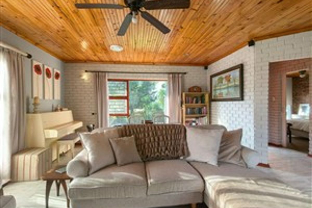 A cozy living room featuring a distinctive timber-paneled ceiling, white painted brick walls, and large windows providing natural light and garden views.