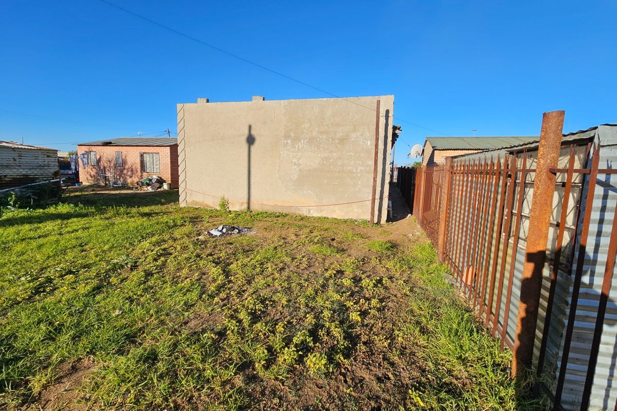 A view of the rear exterior area showing an unpainted concrete structure, rusted metal perimeter fencing, and an unlandscaped yard.