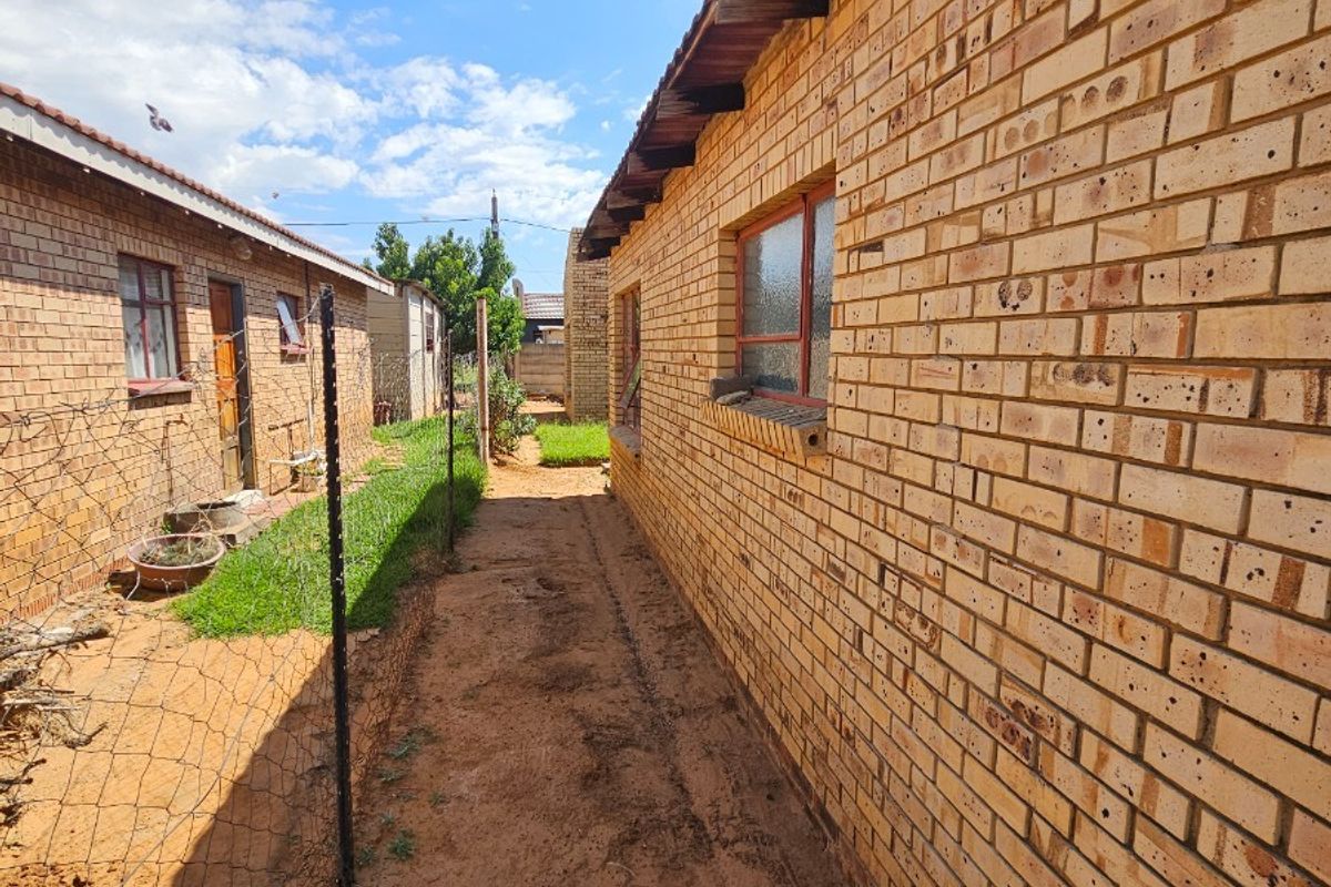 A side exterior view of the property showing brick facade, metal window frames, and an unpaved pathway between two structures.