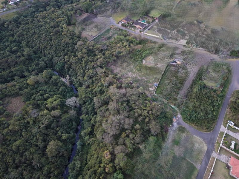 Aerial view of a vacant residential plot featuring natural vegetation, a river boundary, and established road access in a quiet neighborhood setting.