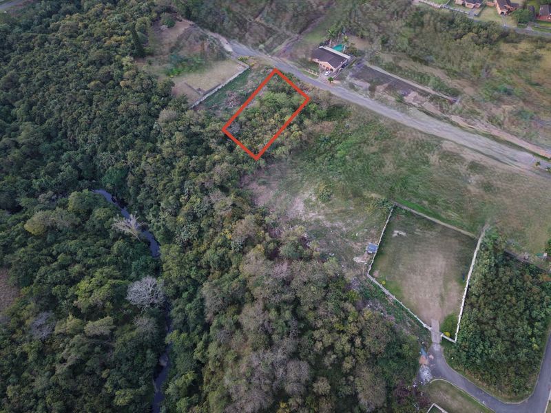 An aerial view of a vacant residential plot highlighted in red, situated in a natural, wooded area near a stream.