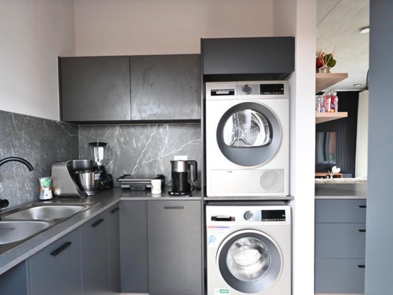 Modern laundry area featuring sleek dark cabinetry, stone-look backsplash, stainless steel sink, and integrated space for a stacked washer and dryer.