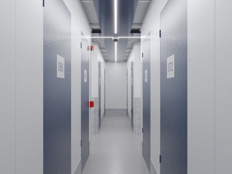 A clean, modern commercial storage hallway featuring numbered unit doors, integrated linear ceiling lighting, and security camera surveillance.
