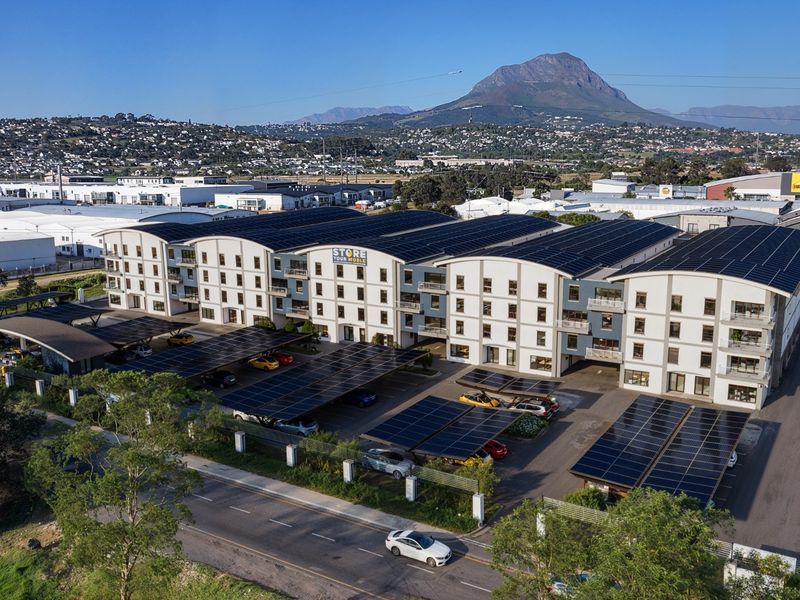Aerial view of a modern commercial complex featuring extensive rooftop and carport solar panel installations, ample parking, and mountain views.