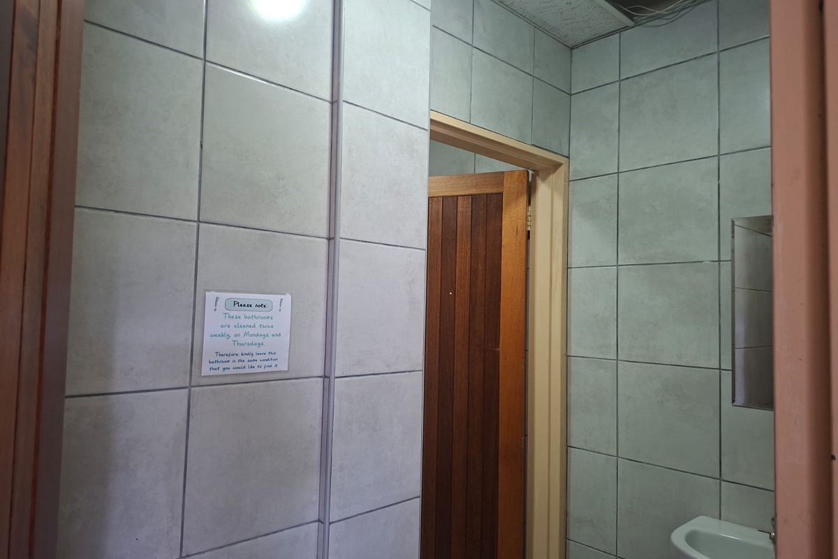 A functional office bathroom featuring tiled walls, a wall-mounted basin, and a wooden door, currently showing signs of wear and ceiling damage.