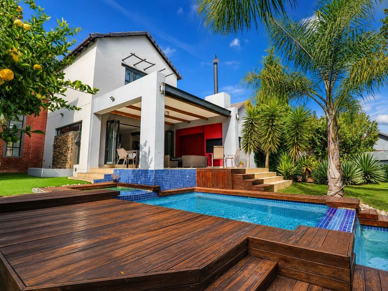 A well-maintained outdoor entertainment area featuring a swimming pool with timber decking, a covered patio with a built-in fireplace, and landscaped garden surroundings.