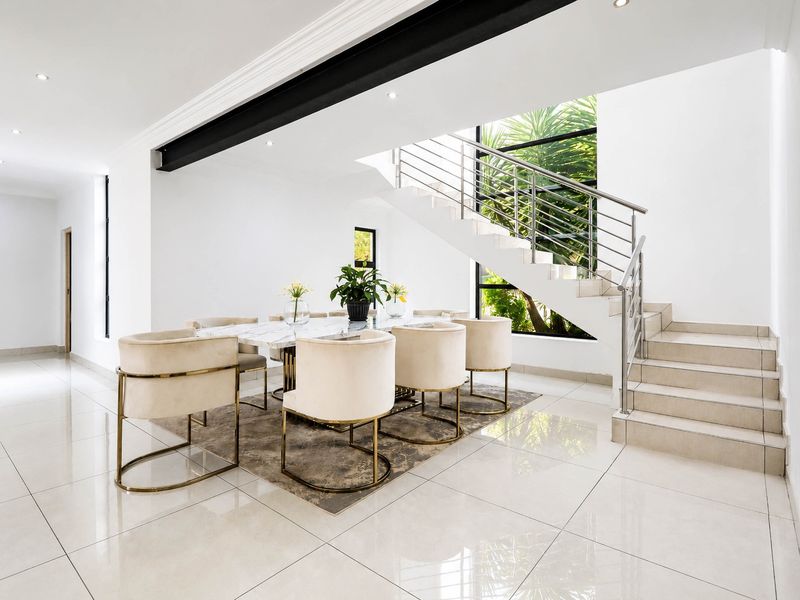 Modern dining area featuring polished tiled floors, an exposed structural beam, and a sleek staircase with a stainless steel balustrade overlooking a garden view.