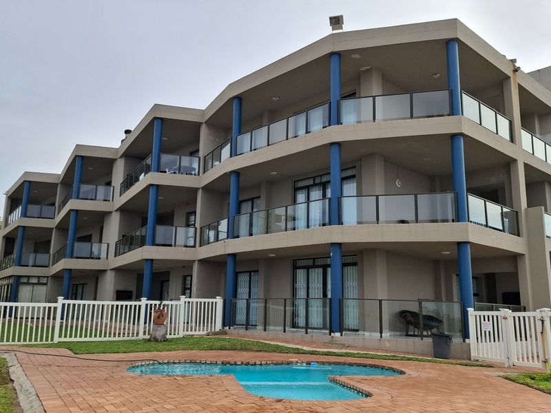 Exterior view of a multi-story apartment complex featuring private balconies with glass balustrades, blue structural columns, and a communal swimming pool area.