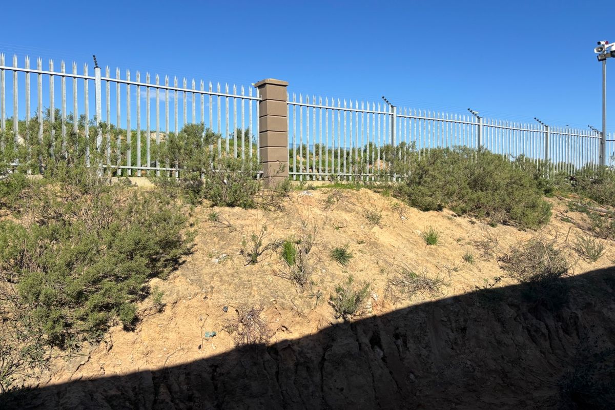 Rear boundary featuring steel palisade fencing with integrated electric security wires and a mounted surveillance camera overlooking an unlandscaped embankment.