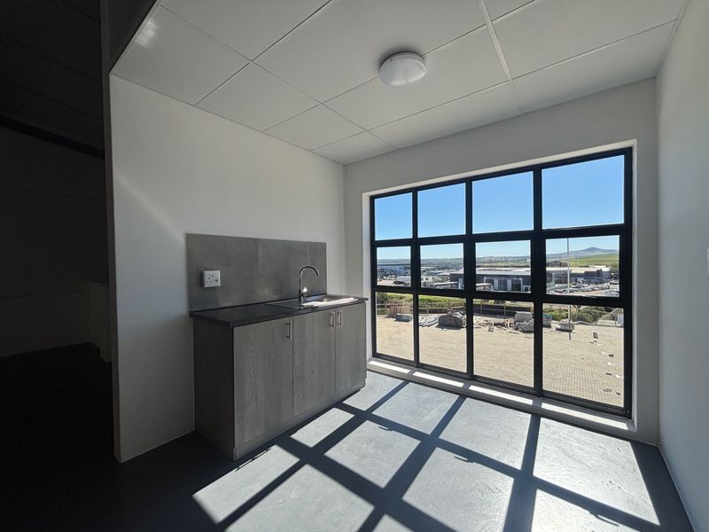 A clean, modern kitchenette featuring grey cabinetry, a stainless steel sink, and a large window offering an industrial park view.