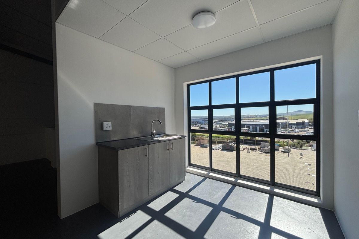 A clean, modern kitchenette featuring grey cabinetry, a stainless steel sink, and a large window offering an industrial park view.