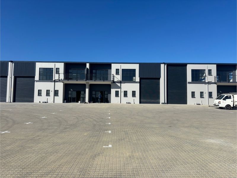 Modern industrial complex featuring multiple units with roller shutter doors, upper-level balconies, and a spacious paved parking forecourt.