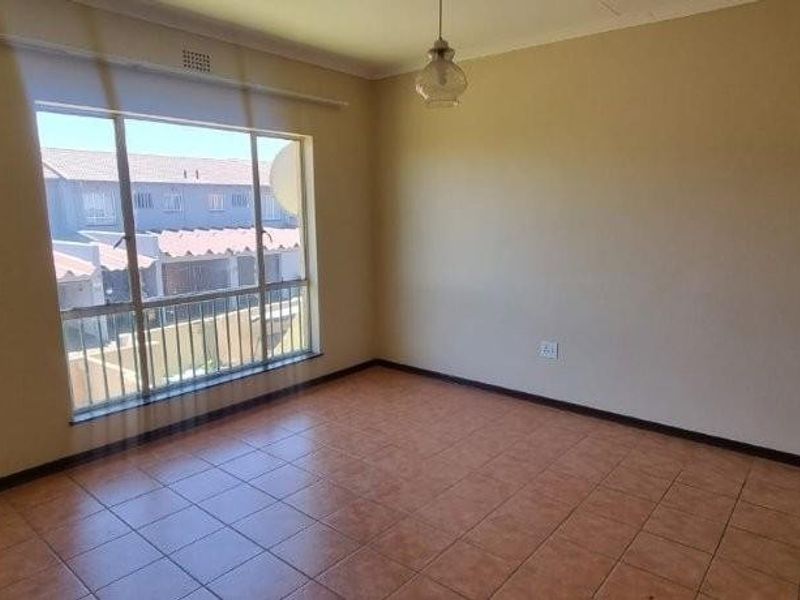 A bright, empty living room featuring tiled flooring, neutral walls, and a large window providing natural light and a view of the complex.