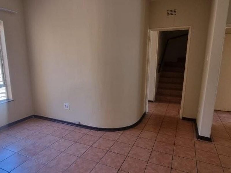 An empty living area featuring tiled flooring, a curved wall detail, and large windows with security bars, leading to a hallway and staircase.