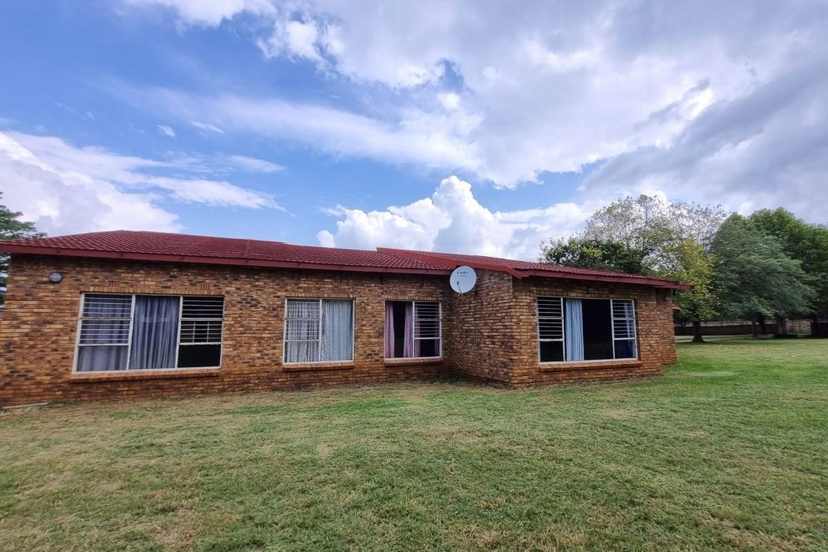 A rear view of a single-story face-brick farmhouse featuring a tiled roof, large windows with security bars, and a spacious surrounding lawn.