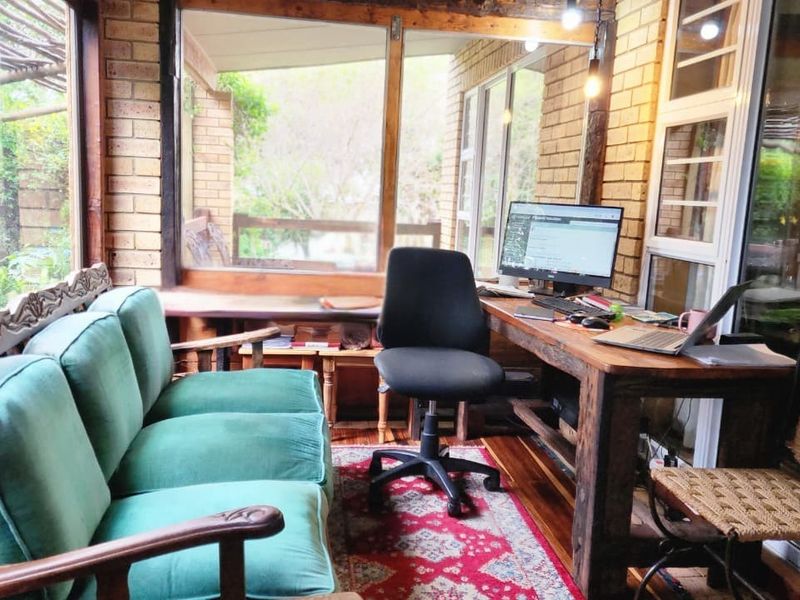 A sun-drenched home office or sunroom featuring exposed brick walls, large windows, and timber flooring, offering a cozy and functional workspace.