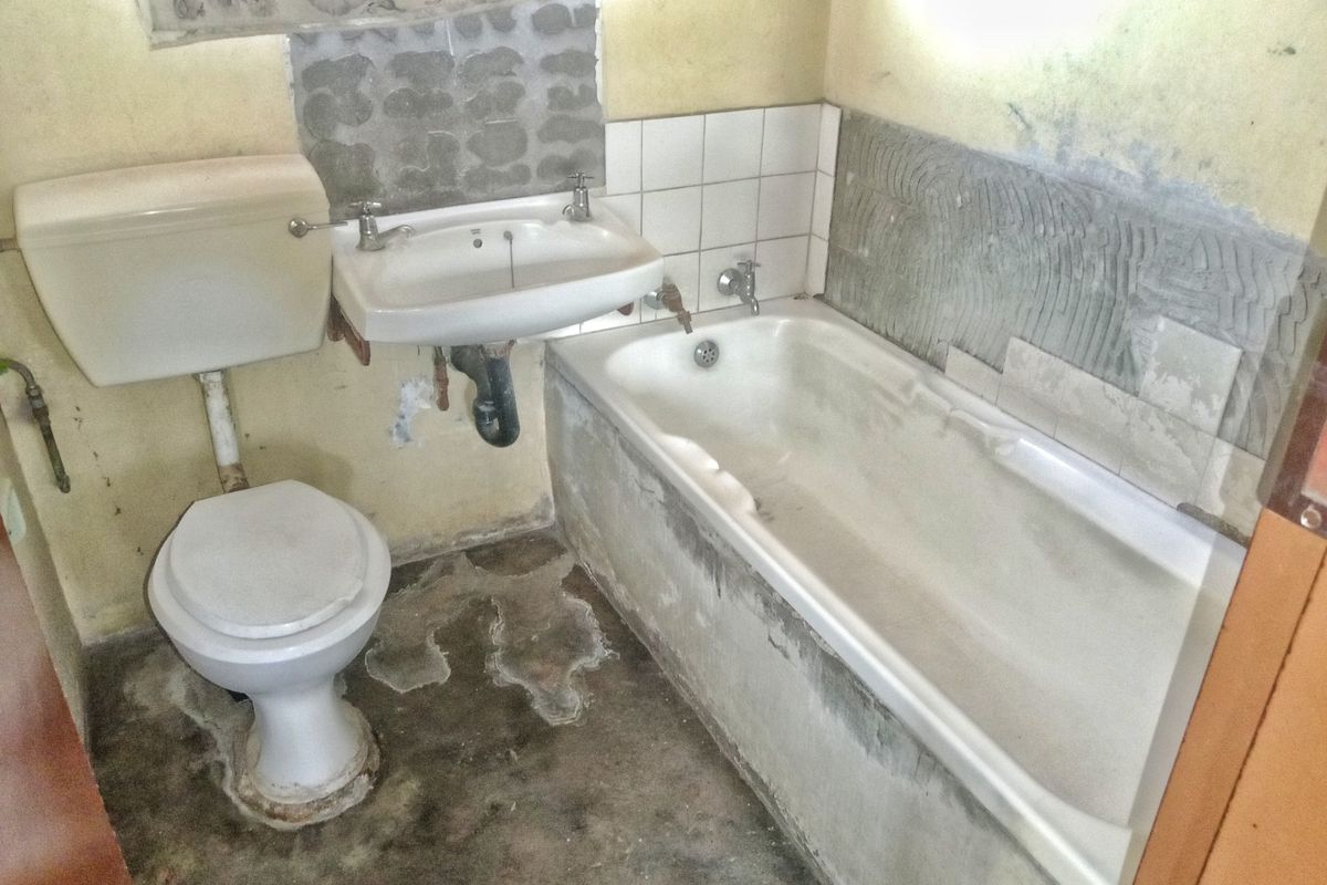 A bathroom in need of significant renovation, featuring a bathtub, pedestal basin, and toilet with unfinished wall tiling and exposed surfaces.