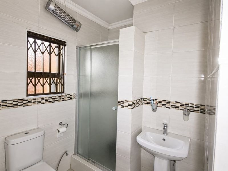 A functional bathroom featuring a pedestal basin, a glass-enclosed shower, and a toilet, finished with neutral wall tiles and a security-fitted window.