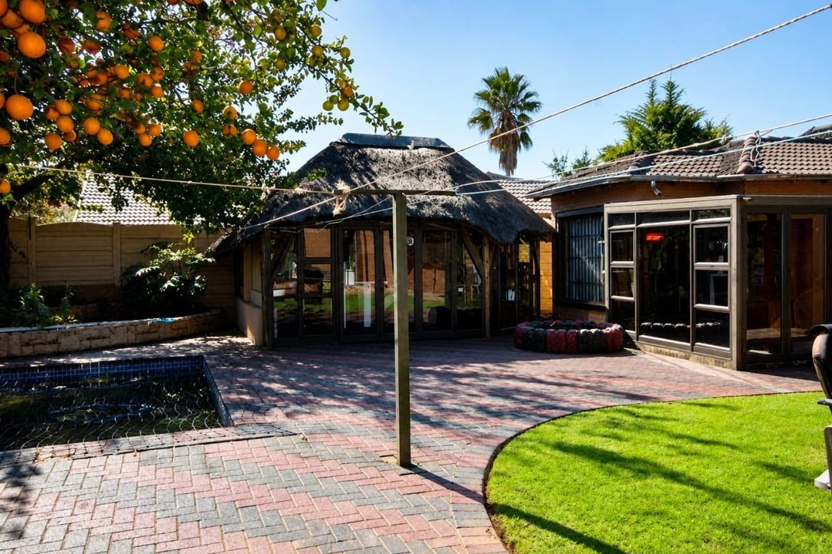 Rear garden area featuring a paved patio, swimming pool, and a thatched roof lapa structure, surrounded by established trees and boundary walls.