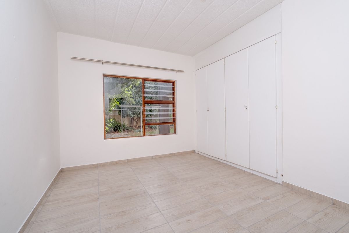 A bright, empty bedroom featuring light wood-look floor tiles, built-in storage cupboards, and a window overlooking the garden area.