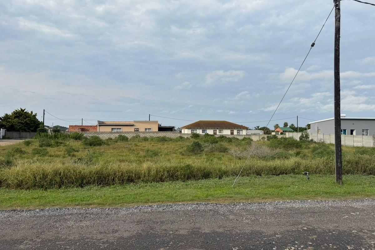 A vacant residential plot of land featuring natural grass cover, enclosed by boundary walls, with access from a gravel road.
