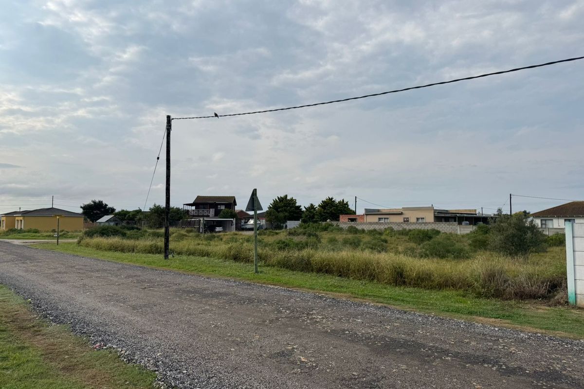 A street-level view of a vacant residential plot featuring a gravel road access and existing utility infrastructure in a suburban setting.