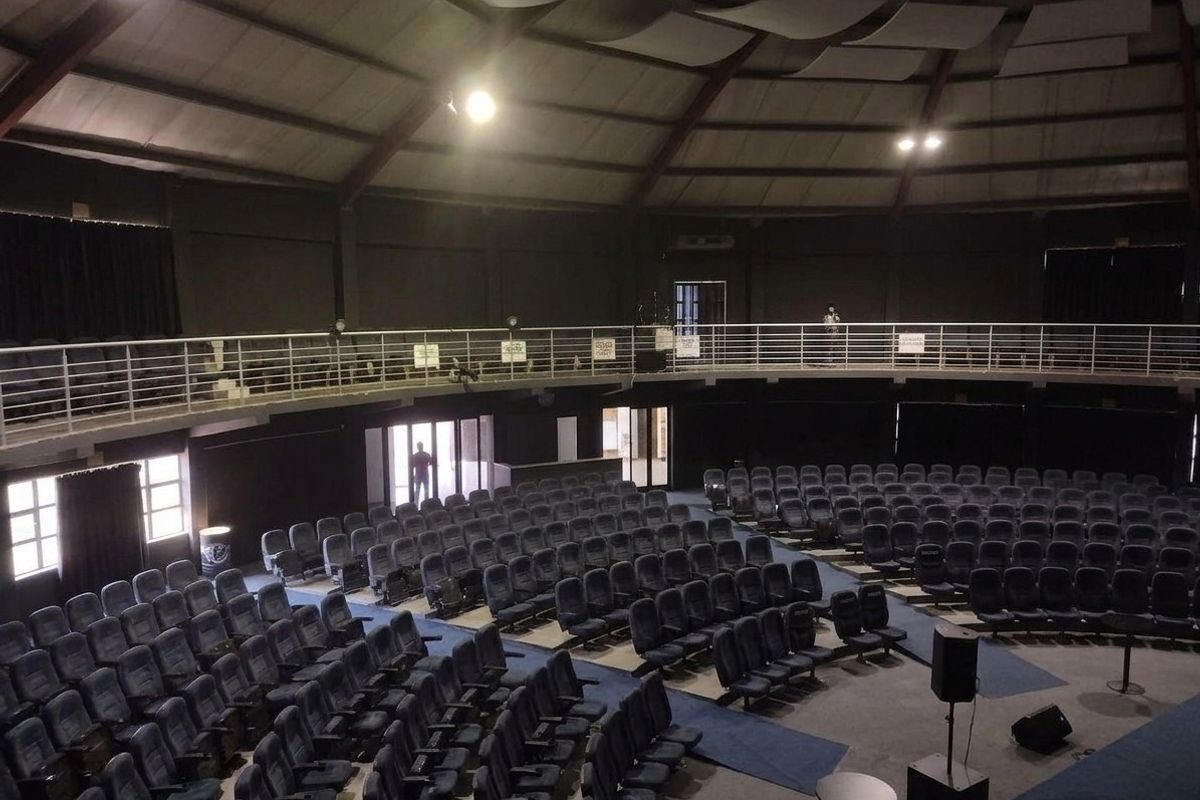 Spacious auditorium or cinema room featuring tiered fixed seating, a mezzanine balcony with metal railings, and exposed steel roof structure.