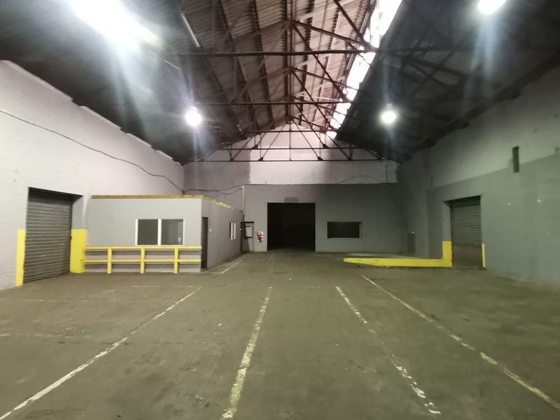 Spacious industrial warehouse featuring high-volume exposed truss ceilings, concrete flooring, multiple roller shutter doors, and an integrated office or utility enclosure.