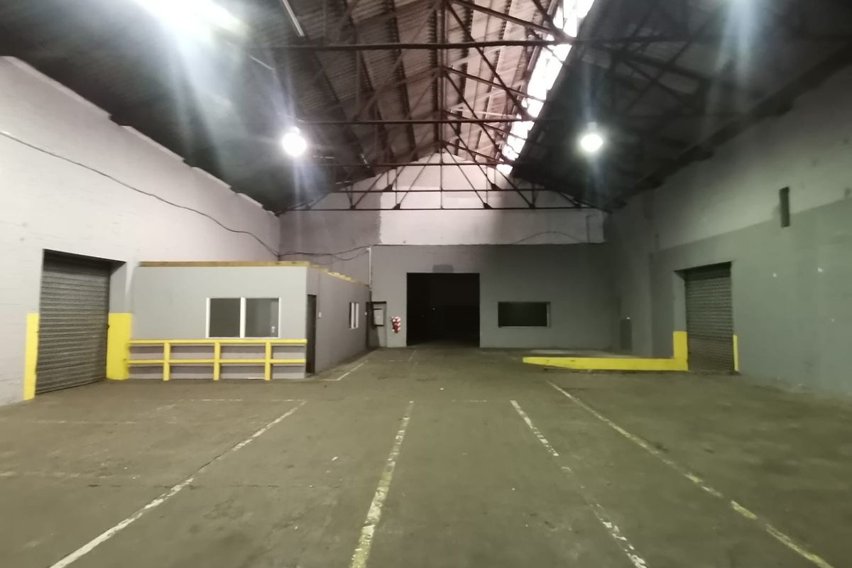 Spacious industrial warehouse featuring high-volume exposed truss ceilings, concrete flooring, multiple roller shutter doors, and an integrated office or utility enclosure.