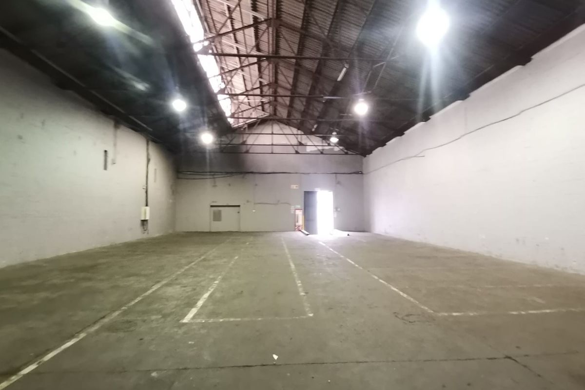 Spacious industrial warehouse featuring high vaulted ceilings, exposed steel trusses, concrete flooring, and ample natural light through roof panels.