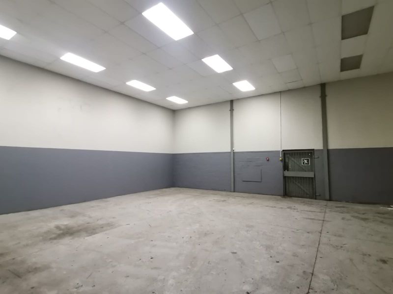 A spacious, empty industrial warehouse unit featuring high ceilings with recessed lighting, concrete flooring, and a secure metal access door.