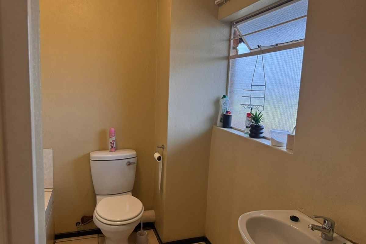 A functional bathroom featuring a standard toilet, washbasin, and tiled flooring, with a louver window providing natural light.