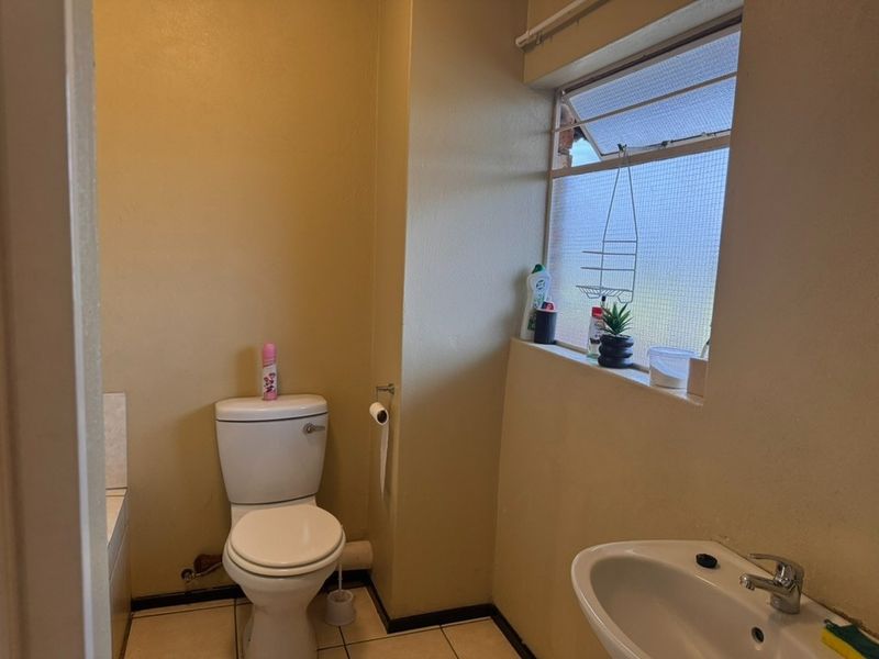 A functional bathroom featuring a standard toilet, washbasin, and tiled flooring, with a louver window providing natural light.