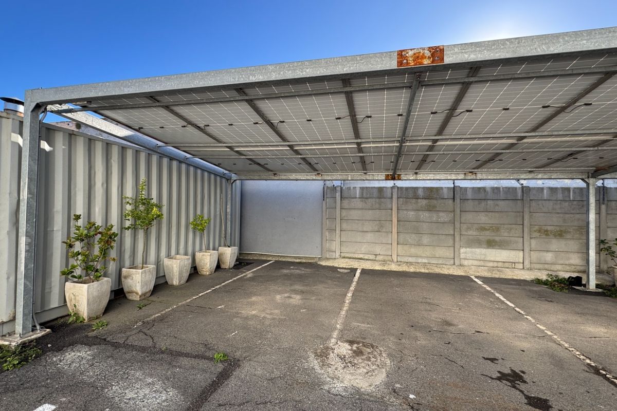 Outdoor parking area featuring a solar panel carport structure, concrete boundary wall, and corrugated metal container wall.